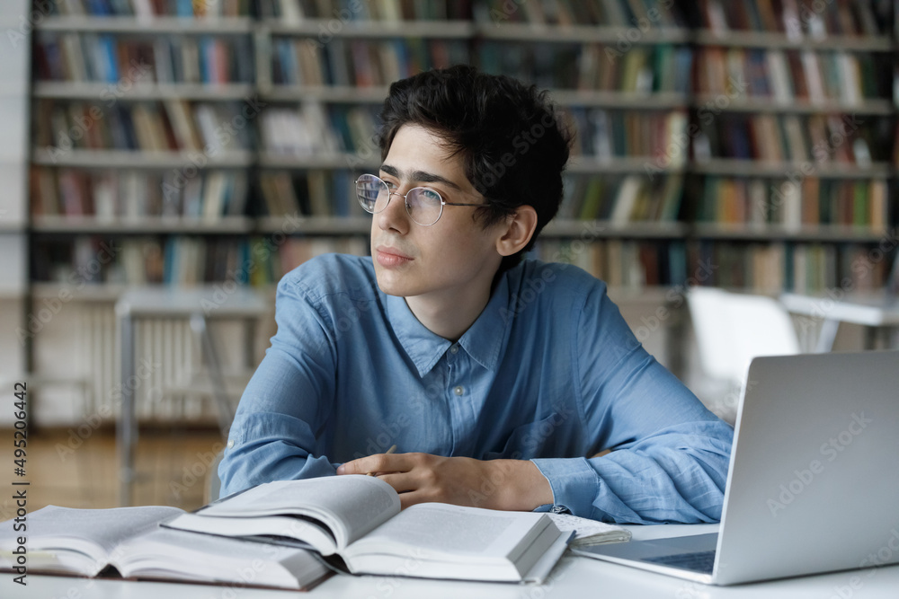 Pensive student guy in glasses staring aside, sit at table in library, distracted from studies, preparation for university admission or college exams looks thoughtful. Education, work on essay concept Pensive student guy in glasses staring aside, sit at table in library, distracted from studies, preparation for university admission or college exams looks thoughtful. Education, work on essay concept