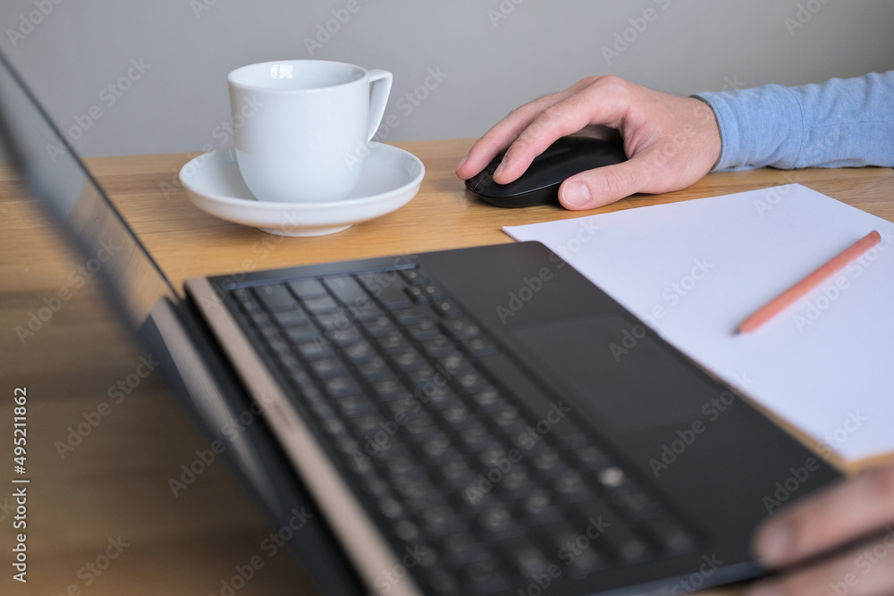 The hand of a man working at a laptop lies on a computer mouse, clicks and scrolls next to a cup of coffee at the workplace on the table in the office closeup The hand of a man working at a laptop lies on a computer mouse, clicks and scrolls next to a cup of coffee at the workplace on the table in the office closeup