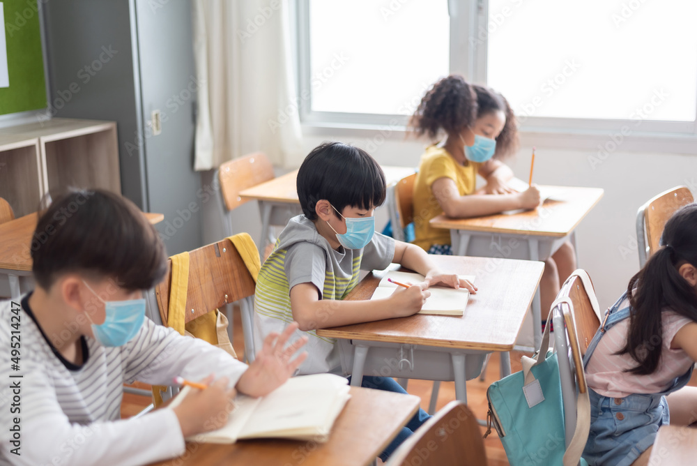 Concentrated Diverse ethnicity small school children wearing protective face mask sitting at the desk in classroom, writing. Concentrated Diverse ethnicity small school children wearing protective face mask sitting at the desk in classroom, writing.