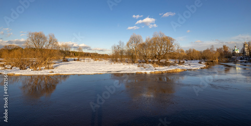 A picturesque landscape, early spring, a river with snow-covered banks, dry grass and bushes. March sunny day by the river. Church in the background. The first thaws, the snow is melting.