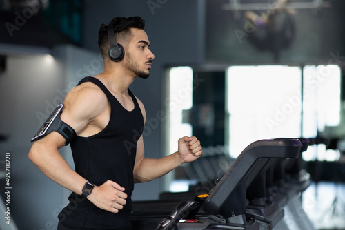 Muscular Arab Male Athlete Wearing Wireless Headphones Running On Treadmill At Gym