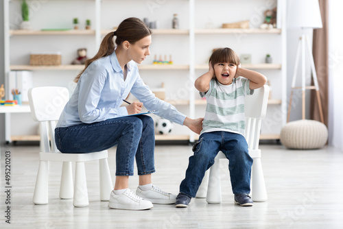 Emotional little boy screaming and covering ears during consultation at psychologist, woman trying to calm down patient