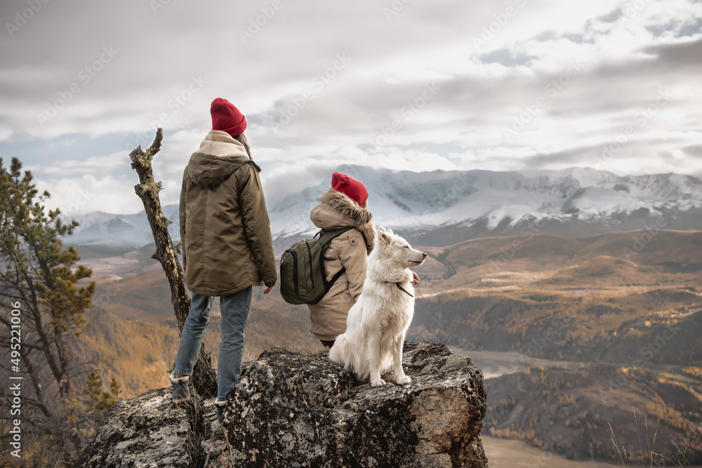 Two female hikers stands at mountain top with white dog and looks at the valley