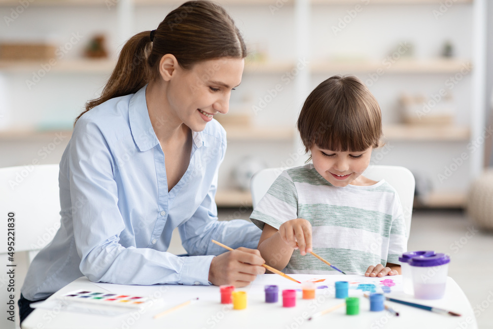 Cute little boy and his young mother drawing together with colorful paints, sitting at classroom and smiling Cute little boy and his young mother drawing together with colorful paints, sitting at classroom and smiling