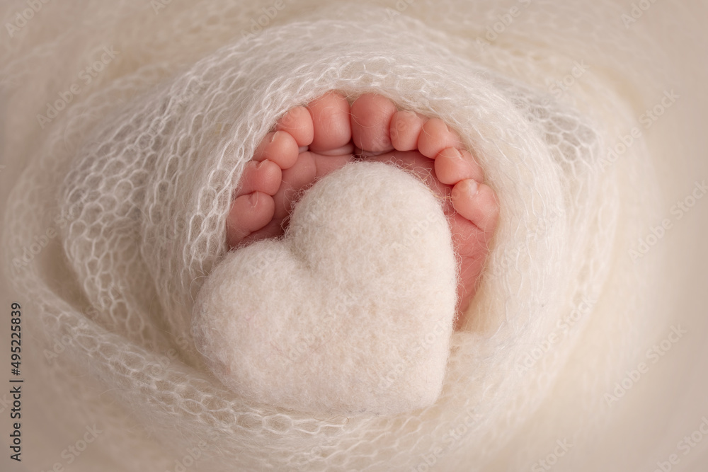 The tiny foot of a newborn baby. Soft feet of a new born in a white wool blanket. Close up of toes, heels and feet of a newborn. Knitted white heart in the legs of a baby. Macro photography. The tiny foot of a newborn baby. Soft feet of a new born in a white wool blanket. Close up of toes, heels and feet of a newborn. Knitted white heart in the legs of a baby. Macro photography.