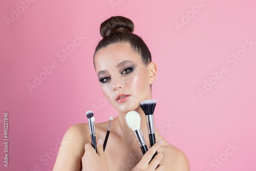 Makeup of a girl's face close-up in the studio on a pink background in the hands of a brush and paint. A young girl does makeup in the studio, paints eyelashes, eyebrows, lips look in the mirror.