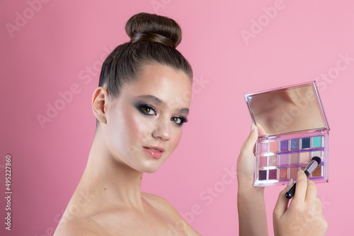 Makeup of a girl's face close-up in the studio on a pink background in the hands of a brush and paint. A young girl does makeup in the studio, paints eyelashes, eyebrows, lips look in the mirror.