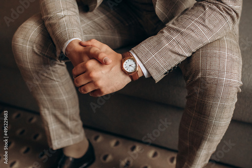Man in a stylish suit with a wristwatch sits and holds his hands together. Groom getting ready in the morning before wedding ceremony. Man sit 