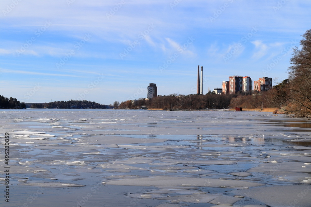 Winter day with ice on the lake. Small town with large chimney in the background. Mälaren, Hässelby, Stockholm, Sweden, Europe.