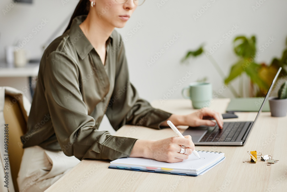 Side view portrait of female business owner writing in notebook while using computer in minimal office interior Side view portrait of female business owner writing in notebook while using computer in minimal office interior