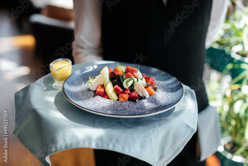 Φωτογραφία Close-up of a young waiter in a stylish uniform carrying an exquisite salad to a client in a beautiful gourmet restaurant