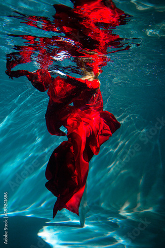 Attractive red-haired young woman swims beautifully underwater in a red dress.