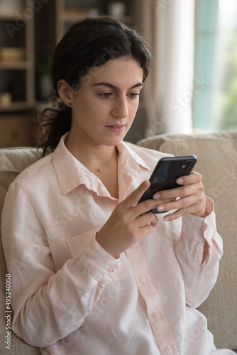 Focused thoughtful beautiful millennial girl typing text message on smartphone, chatting on social media, using payment service, internet banking app on cellphone, sitting on home couch. Vertica shot