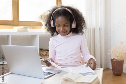 Happy African gen Z school kid in trendy pink headphones studying online from home, sitting at desk with laptop computer, making learning notes, watching online class, smiling. Education on Internet