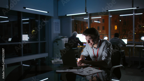 Portrait of happy businessman looking at laptop and laughing working late at night in modern office