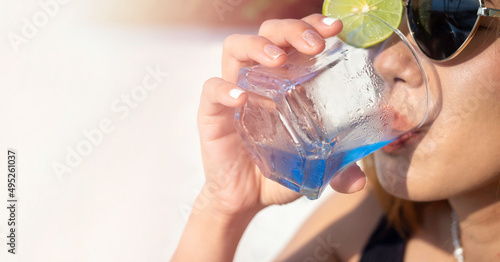 Close-up woman's hand with glitter fashionable nail polish holding blue cocktail drink with lime in the short glass, welcome drink with copy space. Summer banner background. Woman with sunglasses.