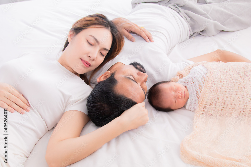 Asian young mother father and newborn sleep on bed together. Parent take a nap with baby because tired nurturing baby. Little infant  deeply sleeping wrapped in thin white cloth with happy and safe.