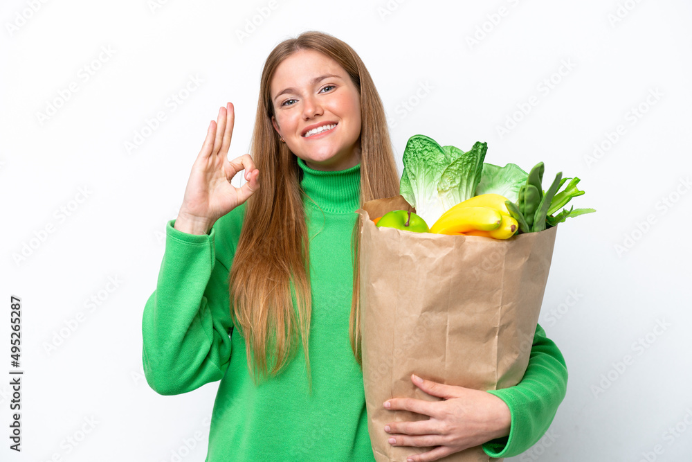 Young caucasian woman holding a grocery shopping bag isolated on white background showing ok sign with fingers Young caucasian woman holding a grocery shopping bag isolated on white background showing ok sign with fingers