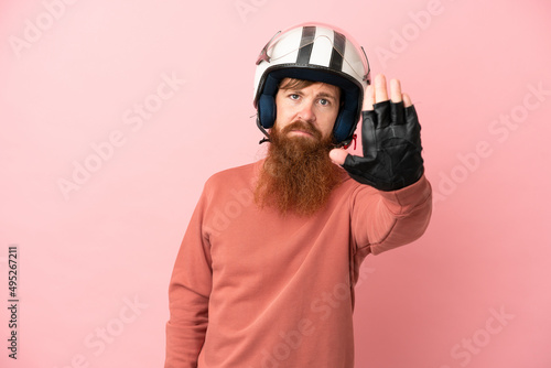 Young reddish caucasian man with a motorcycle helmet isolated on pink background making stop gesture