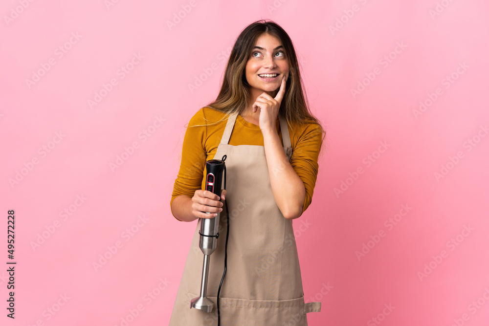 Young chef woman using hand blender isolated on pink background thinking an idea while looking up