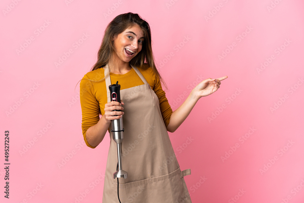 Young chef woman using hand blender isolated on pink background pointing finger to the side and presenting a product