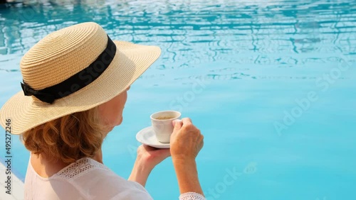 female 50 years old having breakfast by the pool in a straw hat wearing a white dress. woman sitting by the pool with a cup of coffee. good morning and day planning