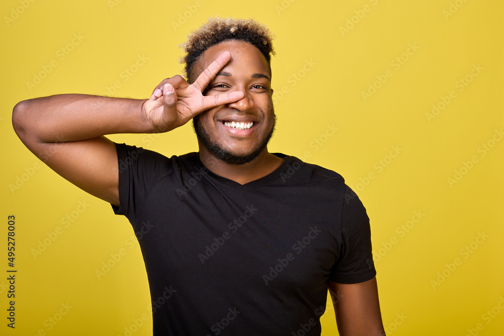 The symbol of peace shows with his right hand a dark-skinned guy in modern everyday clothes on a yellow background. Smiling broadly African-American demonstrates peace and kindness around him The symbol of peace shows with his right hand a dark-skinned guy in modern everyday clothes on a yellow background. Smiling broadly African-American demonstrates peace and kindness around him