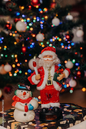 Toy Santa Claus with a bag of gifts in his hands and a snowman on the background of festive Christmas lights. Santa Claus figurine. Snowman figurine. bokeh. Christmas. New Year