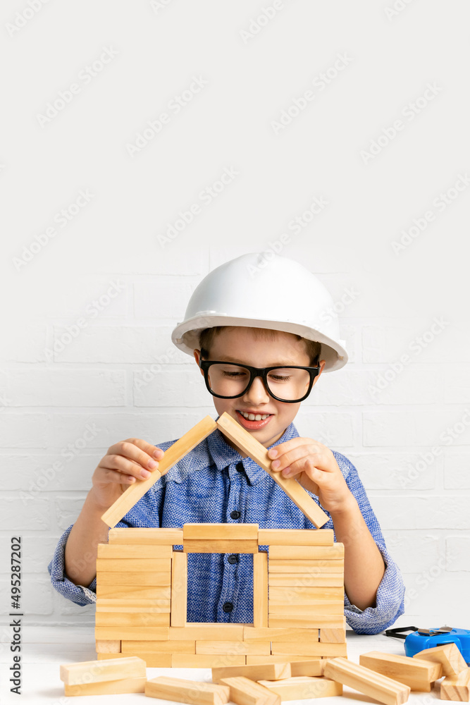 Smiling boy builder in a construction helmet builds a house from wooden blocks. Child engineer play with cubes at home. Mortgage, investment, choice of profession. Copy space. Smiling boy builder in a construction helmet builds a house from wooden blocks. Child engineer play with cubes at home. Mortgage, investment, choice of profession. Copy space.