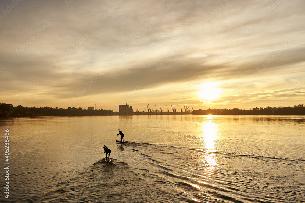 River landscape with silhouette of a two paddle on stand up paddle boarding (SUP) at sunrise on quiet surface of autumn Danube river. Morning training and meditation on the water