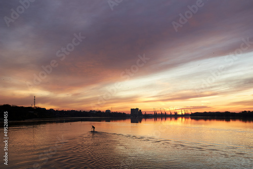 River landscape with silhouette of a teenager boy paddle on stand up paddle boarding  SUP  at sunrise on quiet surface of autumn Danube river. Morning training and meditation on the water