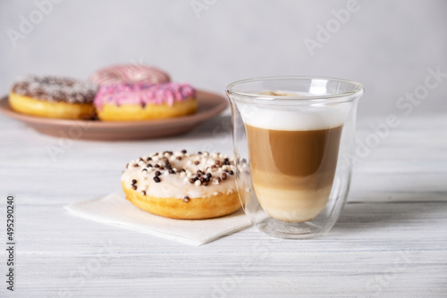 Glazed decorated donuts on plates and coffee latte or cappuccino with milk foam in a heat-resistant glass cup. Selective focus.