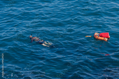 Spearfisherman in wetsuit swims on surface of blue sea water with small orange buoy-boat floating behind him. Hobbies and interests theme.