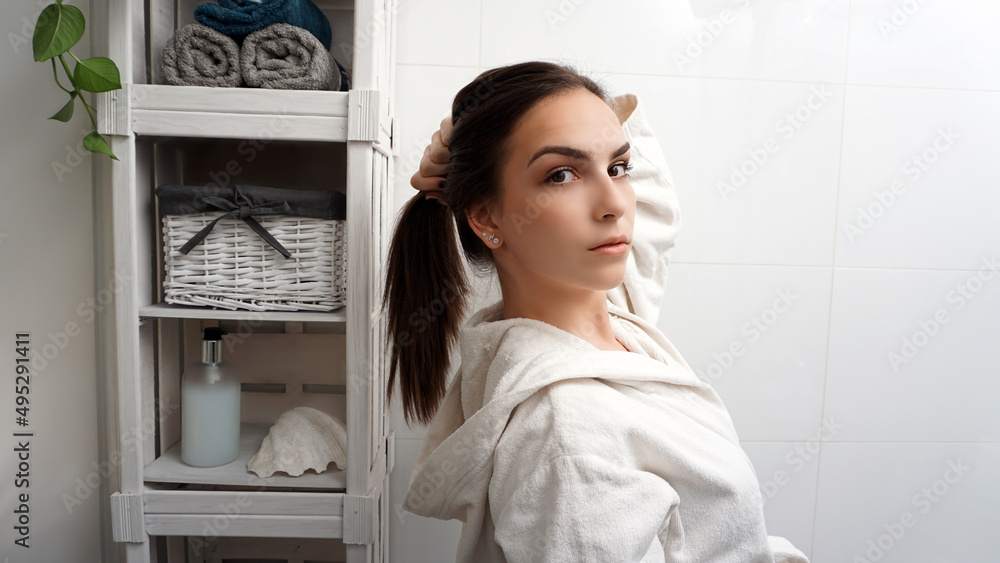 Beautiful young brunette holding her hair in a ponytail and posing in front of the mirror. Girl in bathrobe morning bathroom routine. Happy healthy confident woman. Beautiful young brunette holding her hair in a ponytail and posing in front of the mirror. Girl in bathrobe morning bathroom routine. Happy healthy confident woman.