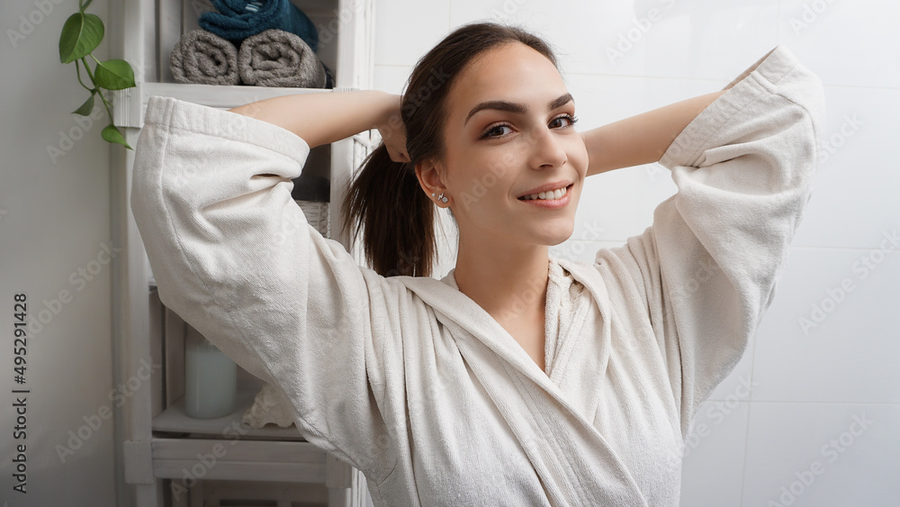 Beautiful young smiling woman in a white bathrobe holding her hair in a ponytail in front of the bathroom mirror. Morning routine. Happy healthy confident life. Beautiful young smiling woman in a white bathrobe holding her hair in a ponytail in front of the bathroom mirror. Morning routine. Happy healthy confident life.
