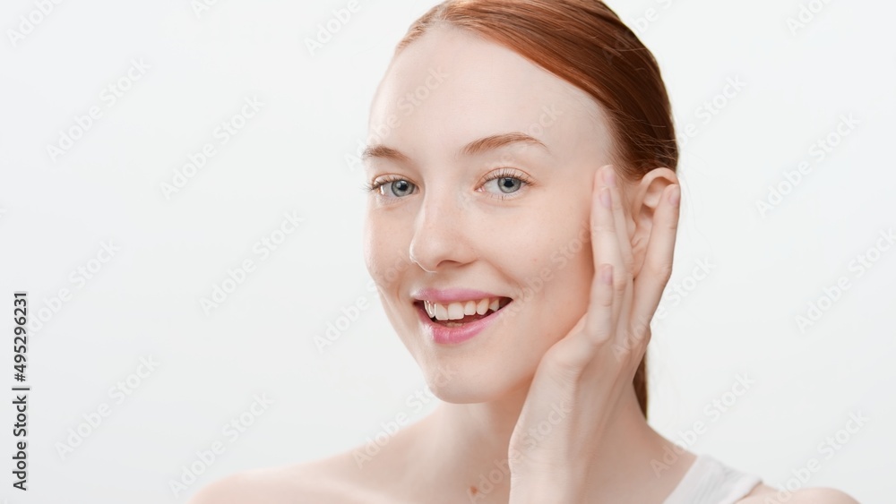 Close-up beauty portrait of young ginger Caucasian woman gently touches her face near temple and cheek smiling for the camera on white background Close-up beauty portrait of young ginger Caucasian woman gently touches her face near temple and cheek smiling for the camera on white background