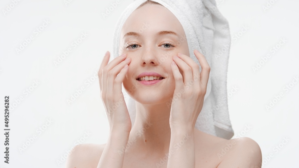 Beautiful slim European young woman with a towel on her hair touches her face gently with fingers of both hands smiling wide for the camera on white background | Face skin care routine concept Beautiful slim European young woman with a towel on her hair touches her face gently with fingers of both hands smiling wide for the camera on white background | Face skin care routine concept