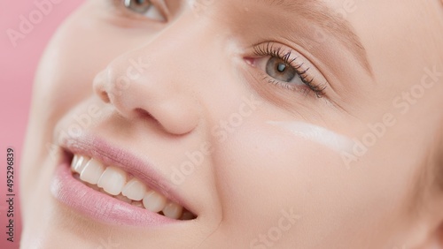 Extreme close-up shot of positive Caucasian woman, beauty model with smear of eye cream under her eye smiling wide on pink background | Eye cream applying shot for face care commercial
