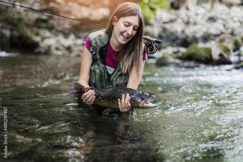 Fototapeta Naklejka Na Ścianę i Meble -  A young woman, who does sports fishing, fishes on a fast mountain river and holds a trout she caught in her hand.