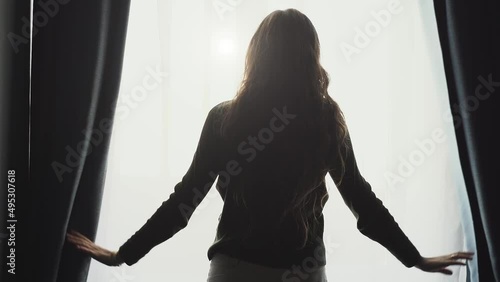 Young woman silhouette open curtains on window to morning light, rear view, slow motion.