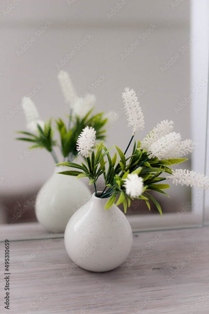 White vase on white in front of a mirror with flowers.