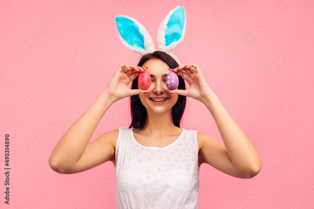 Beautiful and positive brunette woman in bunny ears and with painted Easter eggs pared eyes, easter