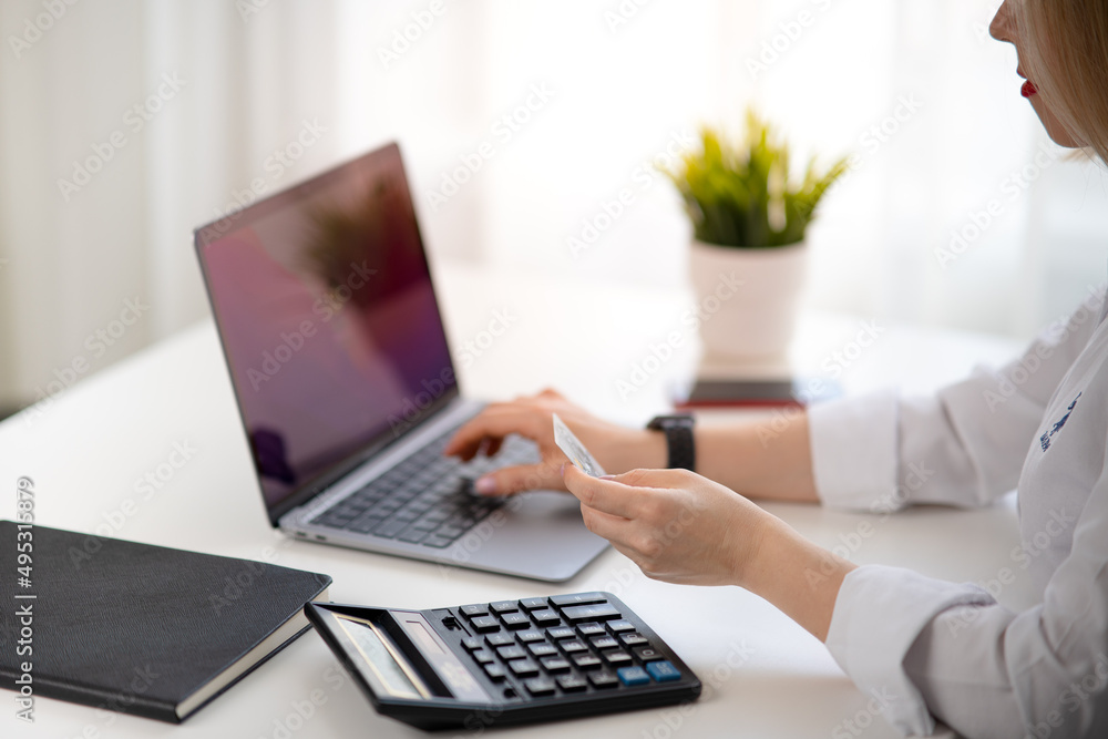 Woman in white shirt holding credit card and entering payment information. Notebook, mobile phone, computer and flower is on the desk. Woman in white shirt holding credit card and entering payment information. Notebook, mobile phone, computer and flower is on the desk.