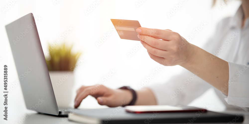 Woman in white shirt holding credit card and entering payment information. Notebook, mobile phone, computer and flower is on the desk.