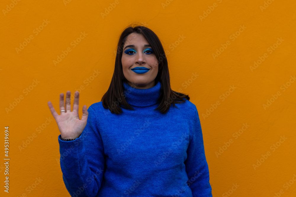 Close up young excited smiling friendly cool caucasian woman 20s with blue make up and blue t-shirt waving hands say hello isolated on yellow color background studio portrait Close up young excited smiling friendly cool caucasian woman 20s with blue make up and blue t-shirt waving hands say hello isolated on yellow color background studio portrait