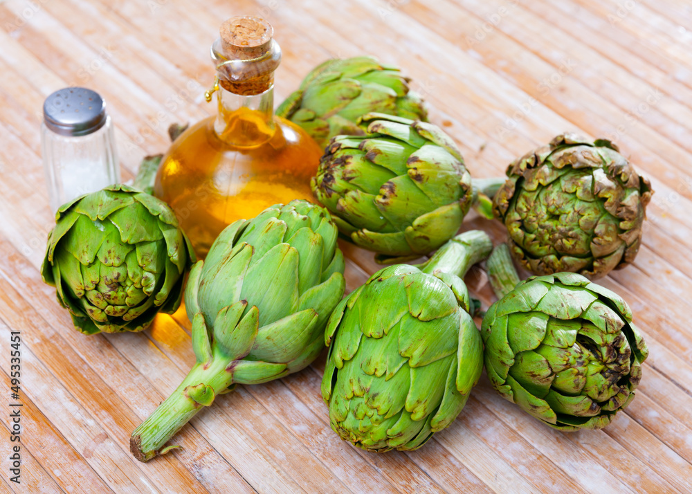 Fototapeta premium Pile of fresh artichokes on wooden table with olive oil in glass decanter and seasonings. Organic vegetarian food concept..