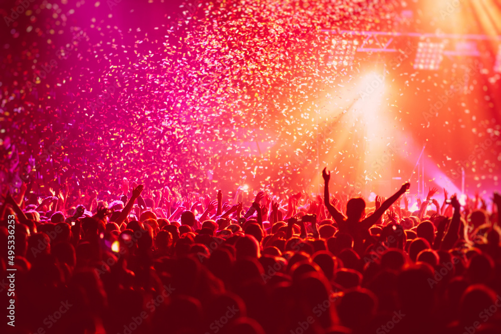 A crowded concert hall with scene stage in red lights, rock show performance, with people silhouette, colourful confetti explosion fired on dance floor air during a concert festival - obrazy, fototapety, plakaty 