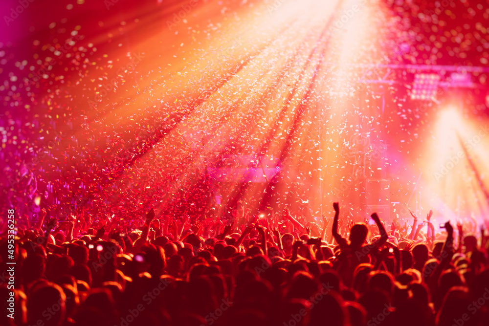 A crowded concert hall with scene stage in red lights, rock show performance, with people silhouette, colourful confetti explosion fired on dance floor air during a concert festival