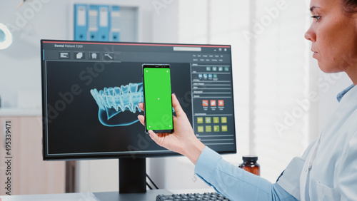Stomatologist vertically holding green screen on mobile phone in dentist office. Dental specialist looking at modern device with isolated background and mockup template for teethcare