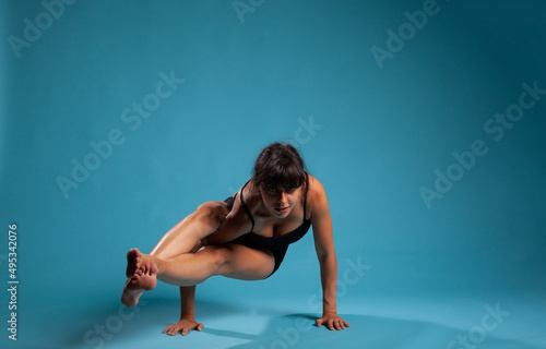 Athletic adult trainer in sportwear working at arm balance practicing yoga position in studio with blue background. Active fit woman in sportwear stretching body muscles during sport workout